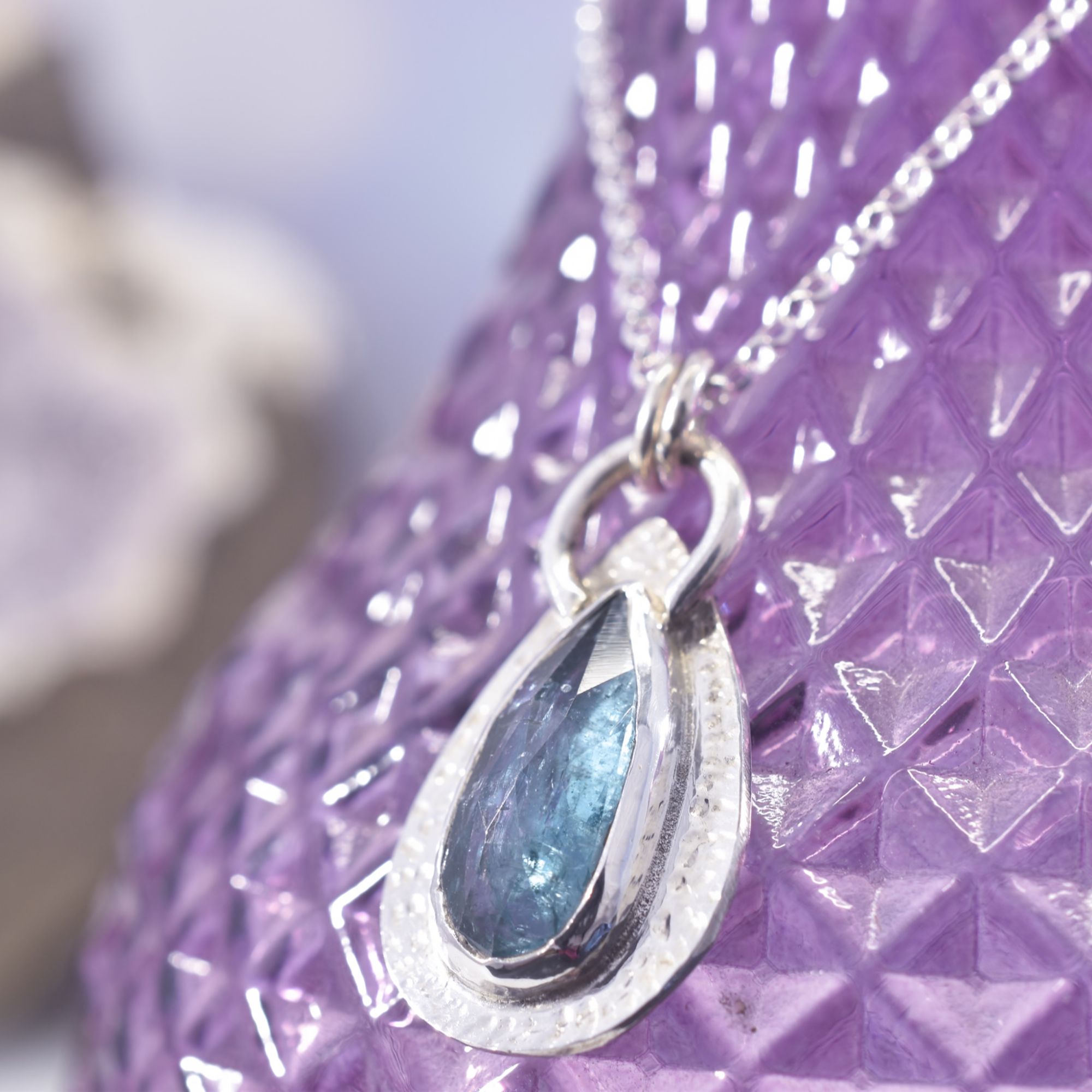 A teardrop of faceted teal ocean kyanite, captured in a textured frame of sterling silver. A cut out to the reverse allows the light to shine through the stone.