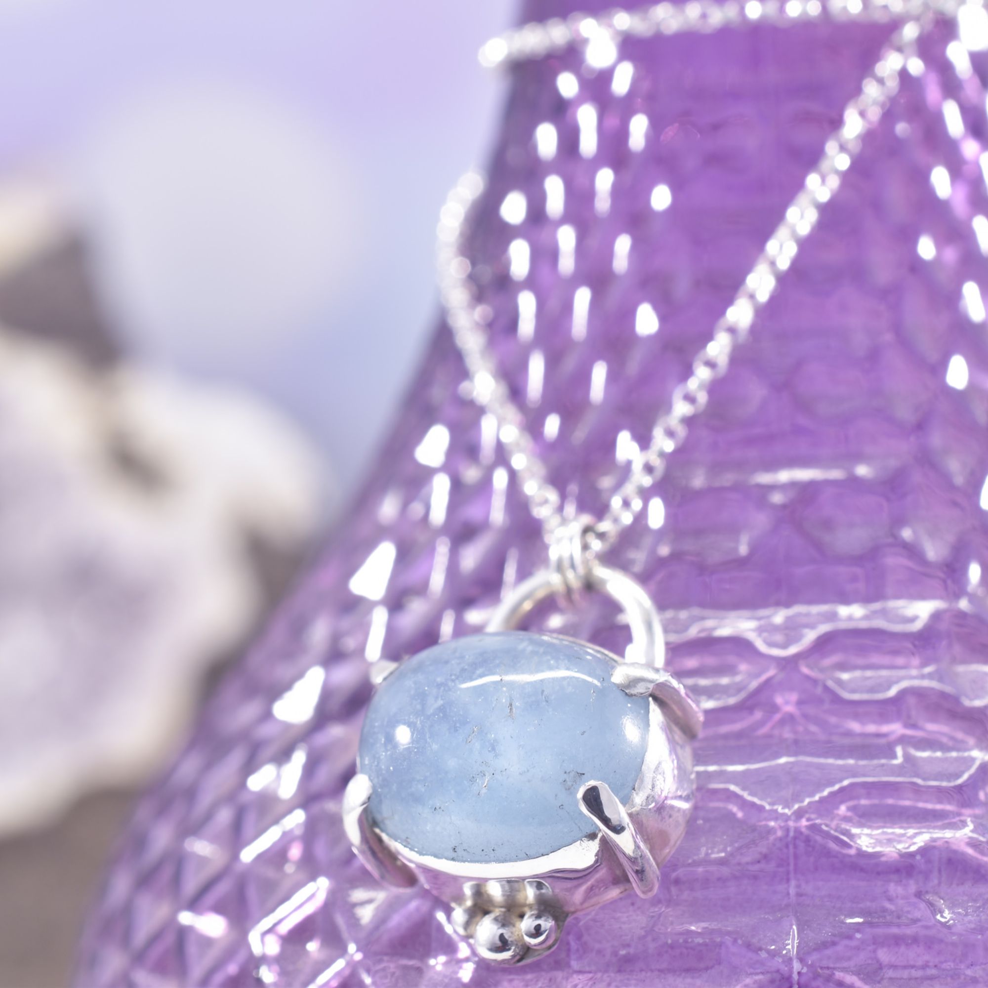 An oval mid blue smooth aquamarine crystal, set horizontally in 4 solid silver claws, with a silver surround, and 3 silver balls to the bottom. The reverse features a hand stamped and sawn make waves design of 3 ocean waves and a cut out to allow the light to shine through the stone.