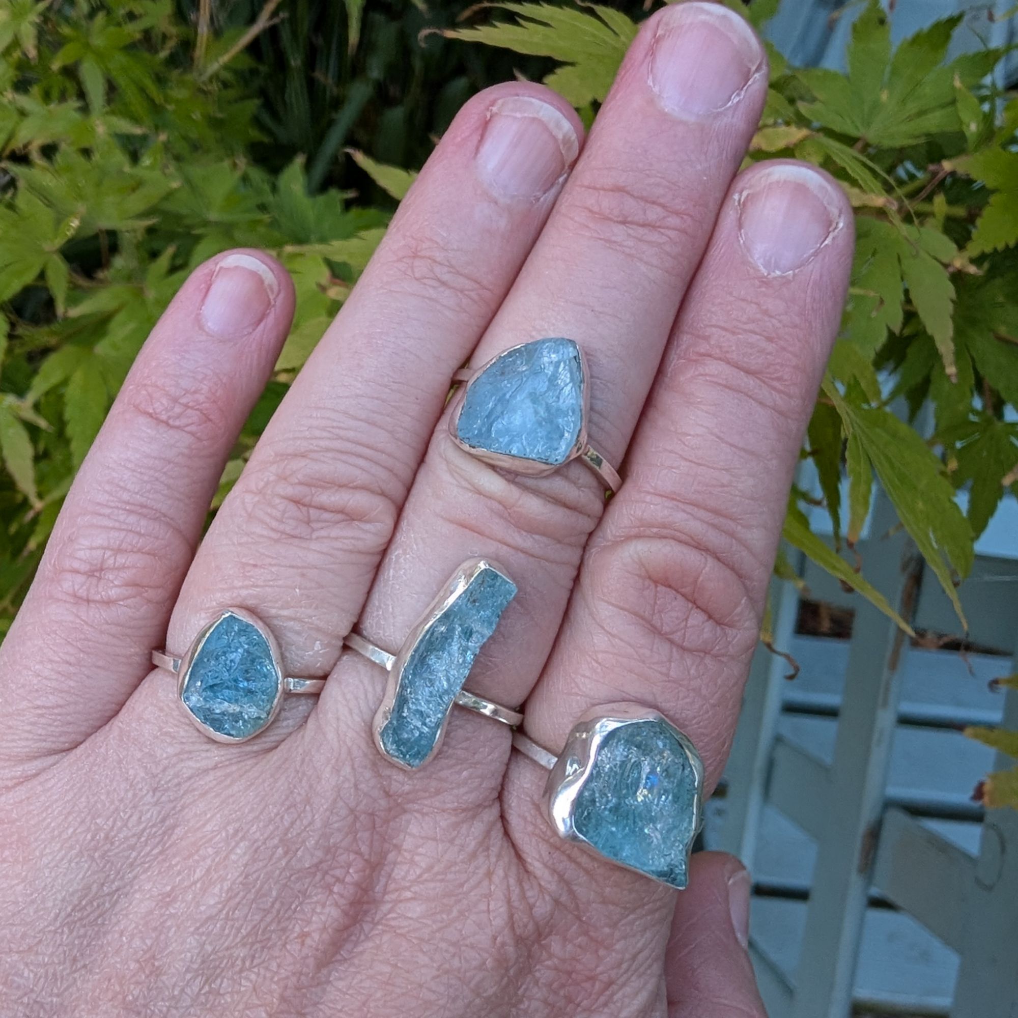 A selection of four individual raw blue aquamarine rings, shown on worn on a hand. Each stone is set in sterling silver and has a square silver ring band.